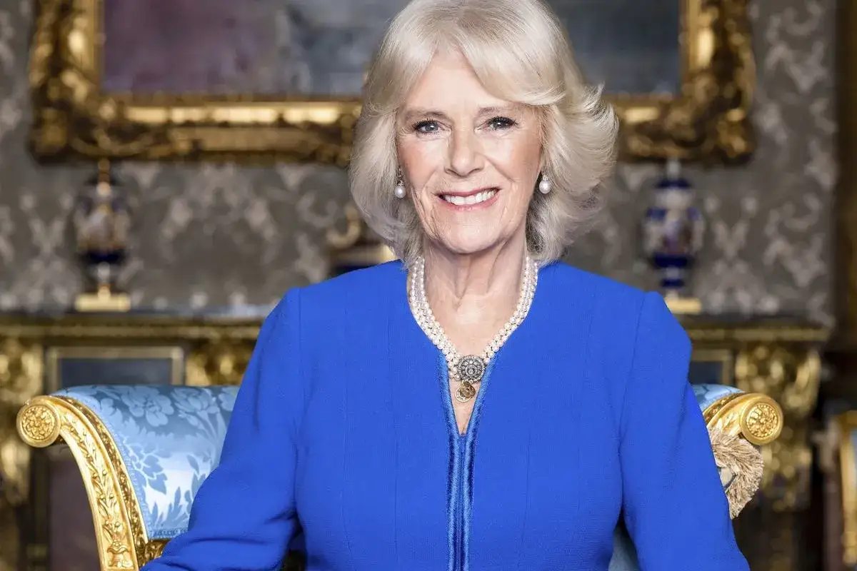 Uśmiechnięta Camilla Parker Bowles w niebieskiej sukience i perłach, siedząca na ozdobnym fotelu.