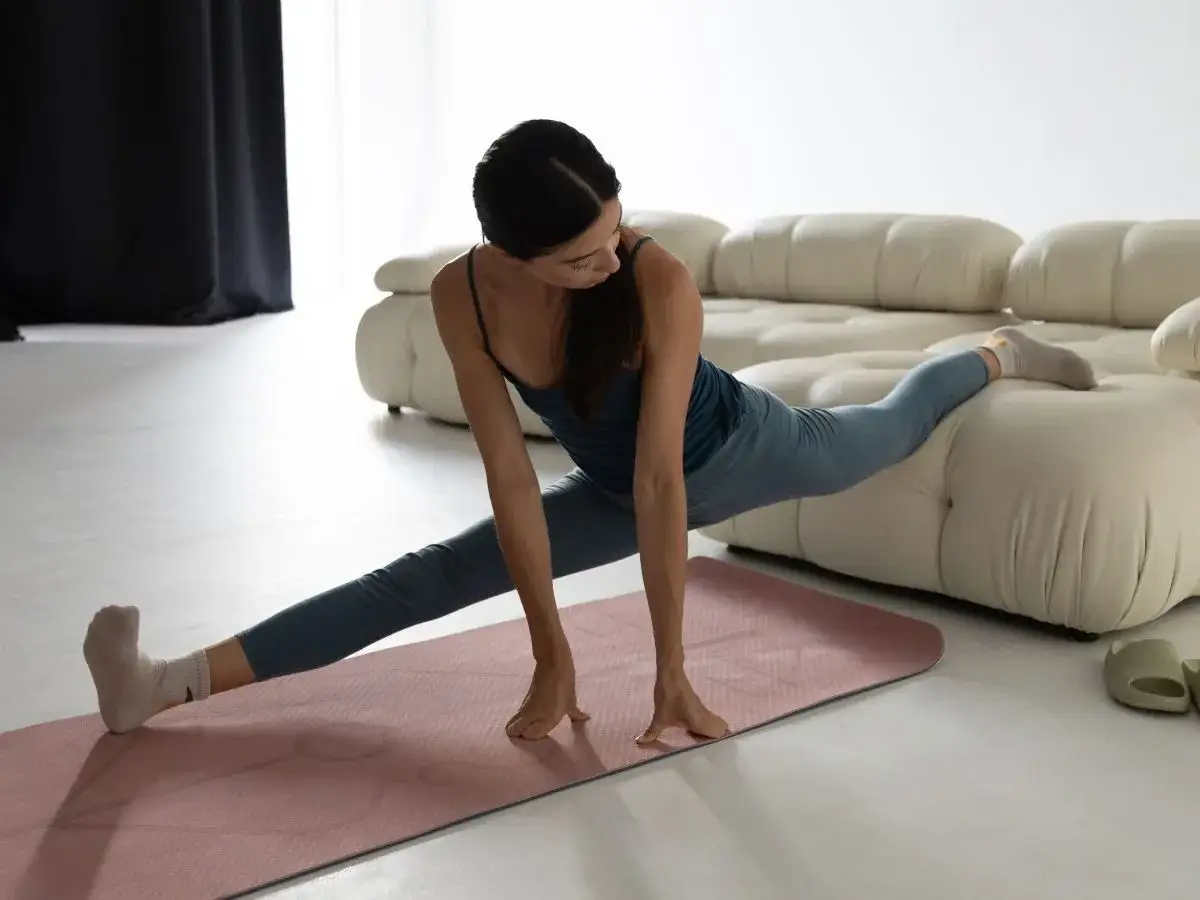 Kobieta wykonuje szpagat na macie, opierając jedną nogę na kanapie.