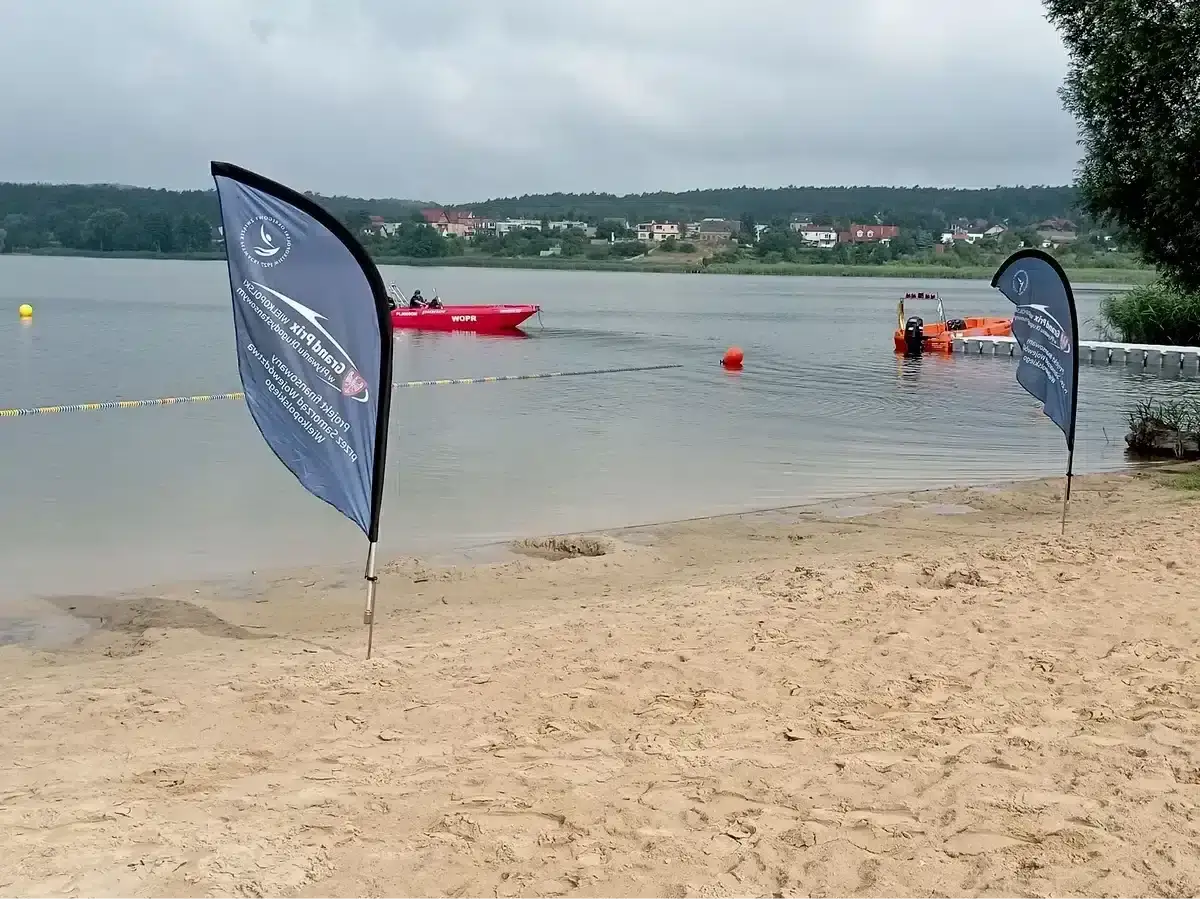 Plaża nad jeziorem, czerwona łódź ratownicza WOPR, pomarańczowa łódź, flagi, budynki na horyzoncie.