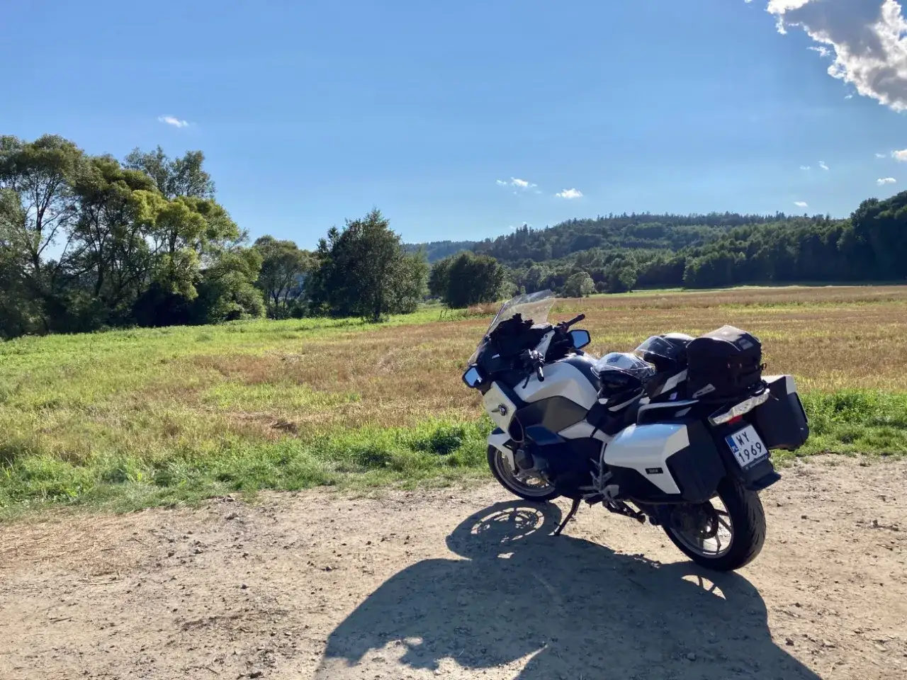 Biały motocykl turystyczny zaparkowany na drodze gruntowej, w tle pole i zalesione wzgórza.