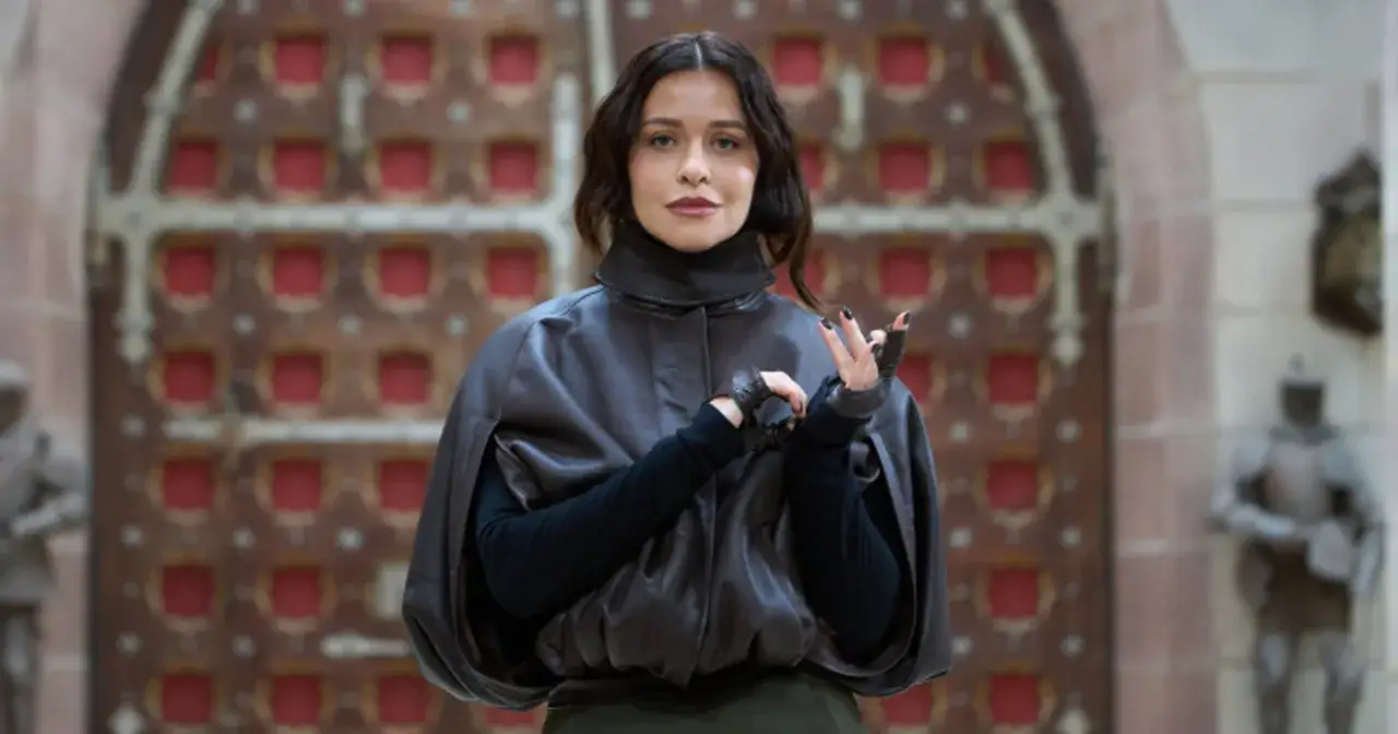 Kobieta w skórzanej pelerynie i rękawiczkach, jakby prowadząca grupę zdrajców, stoi przed ozdobnymi drzwiami.