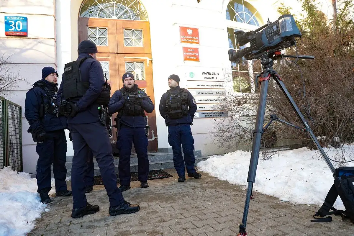 Policjanci stoją przed budynkiem z tabliczkami informacyjnymi, obok stoi kamera na statywie.