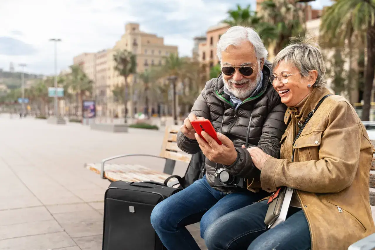 Uśmiechnięta para seniorów na ławce w mieście przegląda telefon. Czyżby sprawdzali, co to roaming, planując kolejną podróż?