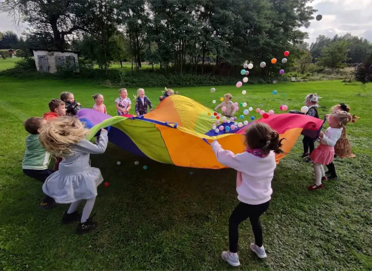 Wesoła animatorka dla dzieci prowadzi zabawę z kolorowym spadochronem i piłeczkami na trawie. Dzieci śmieją się i bawią.