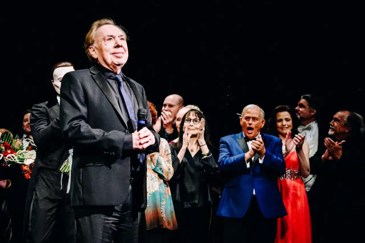 Upiór z Opery na scenie, wita publiczność. Obok niego ludzie w eleganckich strojach, niektórzy klaszczą.