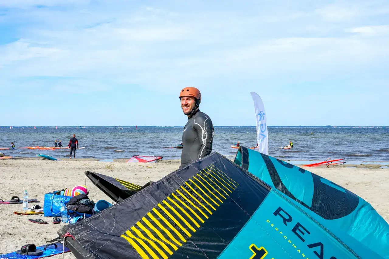 Mężczyzna w kasku i piance na plaży, leżące latawce do kitesurfingu, morze i inni sportowcy.