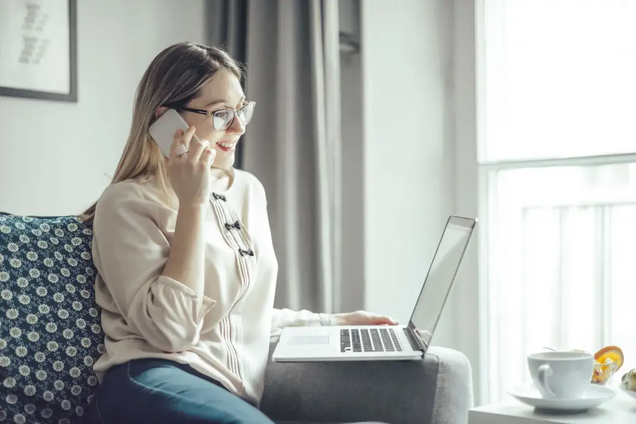 Kobieta w okularach rozmawia przez telefon, trzymając laptopa na kolanach.