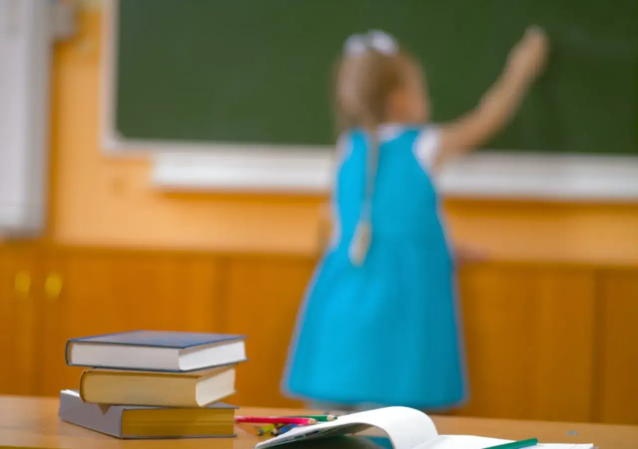 Dziewczynka w niebieskiej sukience pisze kredą po tablicy, a na pierwszym planie leżą książki. Jak to się ma do nauki zdalnej?