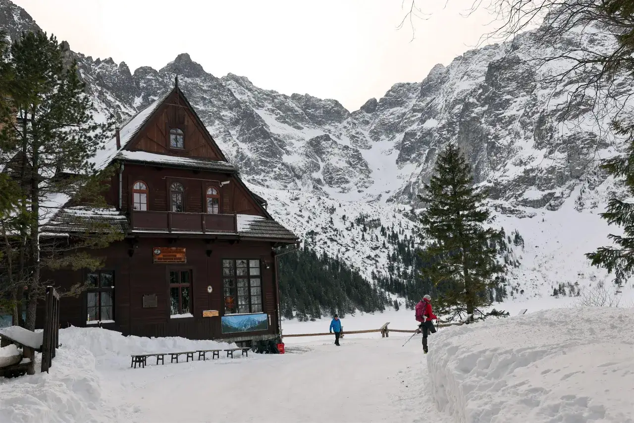 Zimowy krajobraz z drewnianym schroniskiem i ośnieżonymi górami. Turyści wchodzą na teren obiektu. Dowiedz się, ile kosztuje nocleg w schronisku.