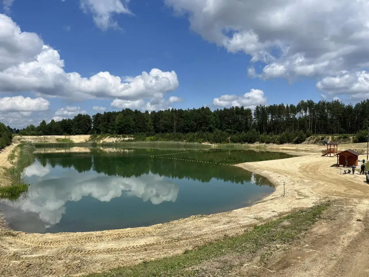 Zbiornik Czarna Sędziszowska z piaszczystą plażą i lasem w tle. Błękitne niebo z białymi chmurami odbija się w spokojnej tafli wody.