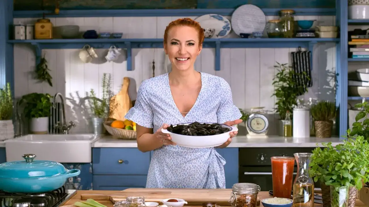 Kobieta w kuchni trzyma miskę z małżami, sugerując, czy dania ma dostęp do morza. W tle widać zioła i naczynia.