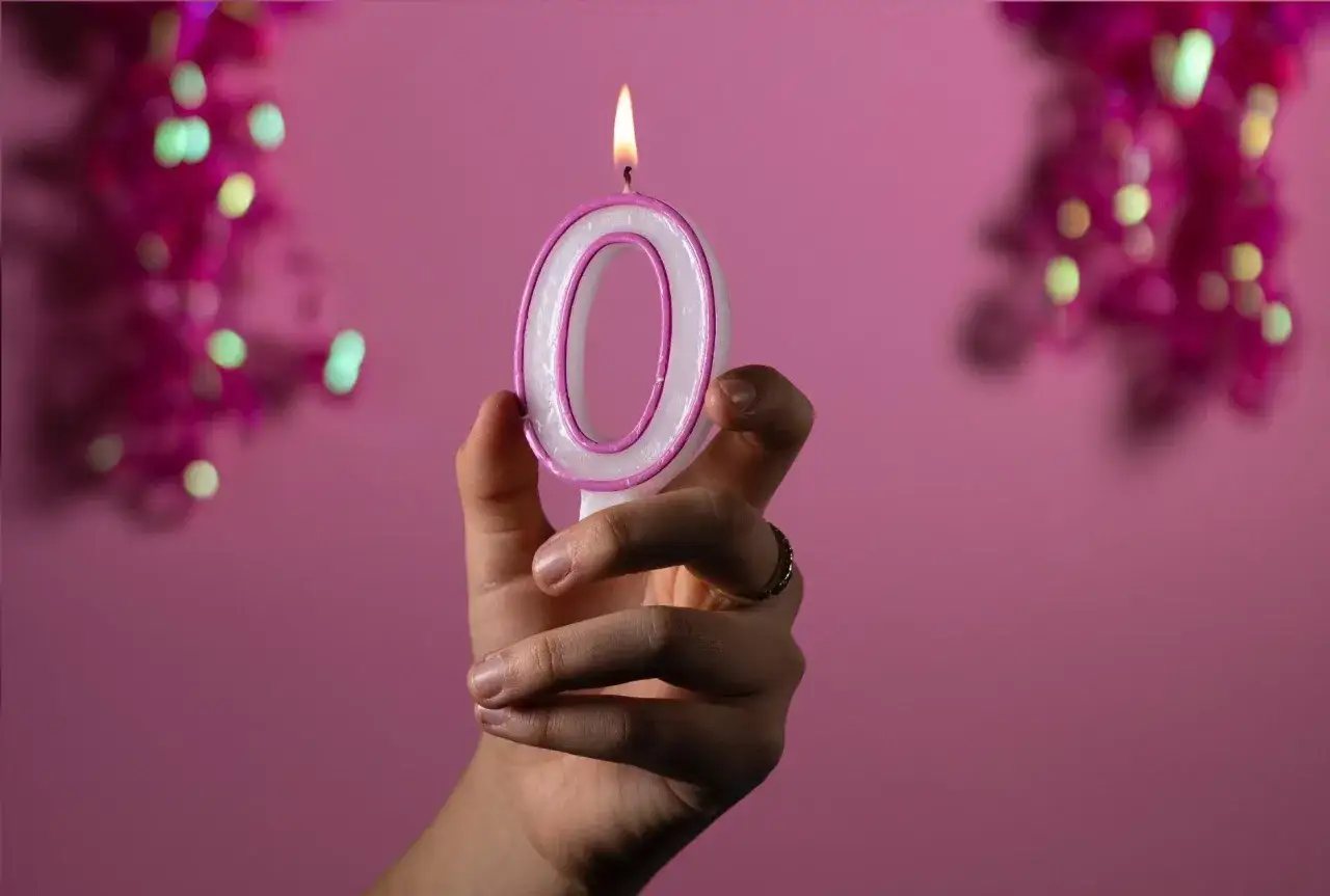 Dłoń trzyma zapaloną świeczkę w kształcie cyfry 0 na różowym tle.