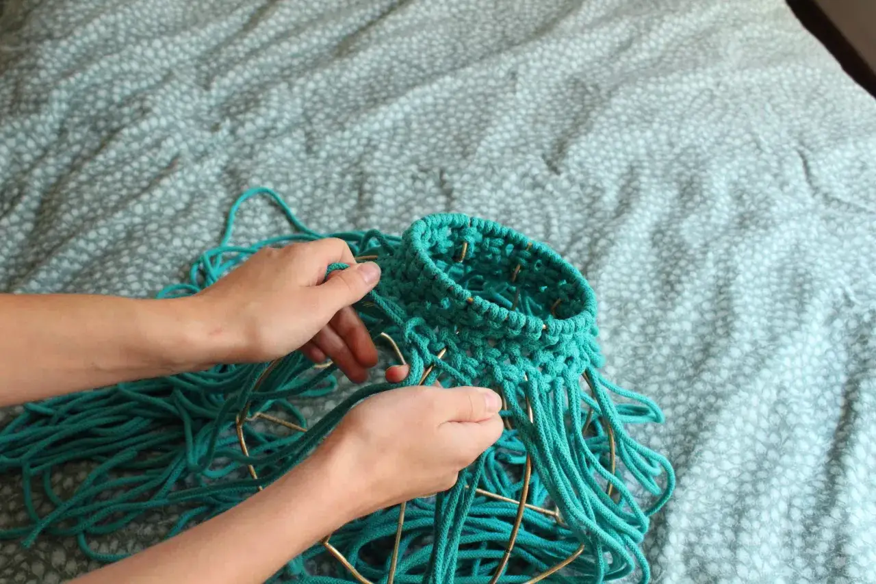 Ręce tworzą makramową osłonę na stary abażur, używając turkusowego sznurka. Oto jak odnowić stary abażur.