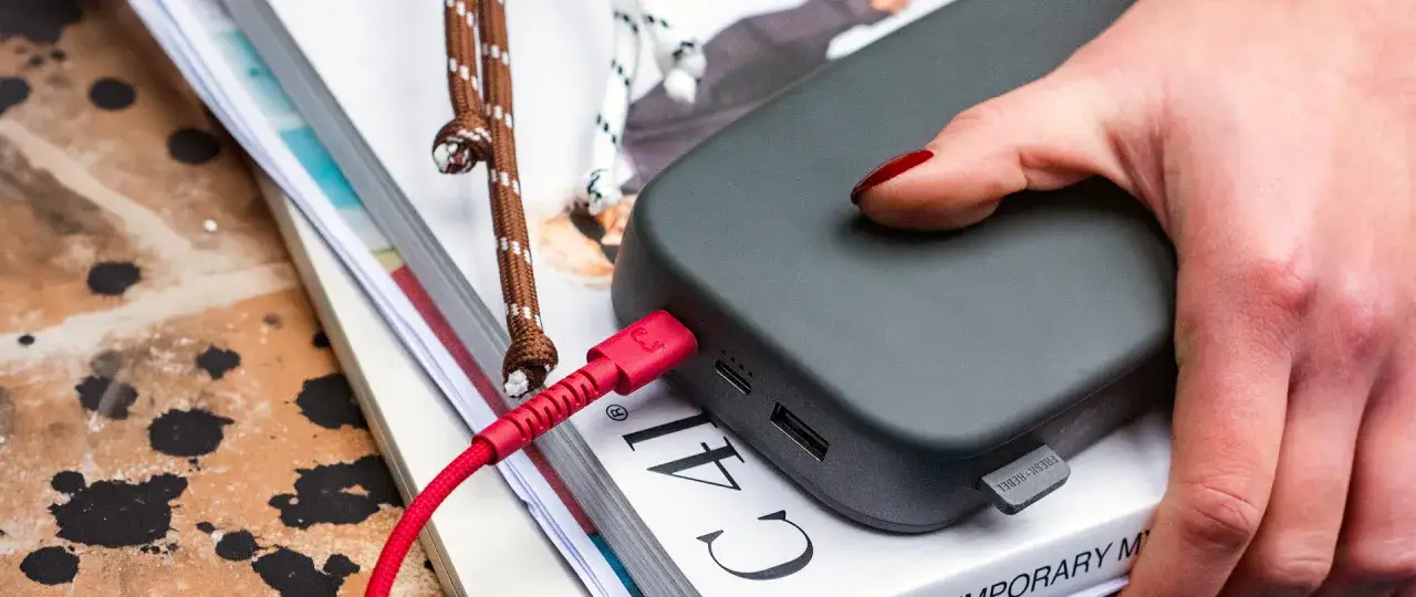 Ciemnoszary power bank 50000 mah na ile starcza? Podłączony czerwonym kablem, leży na stosie książek.
