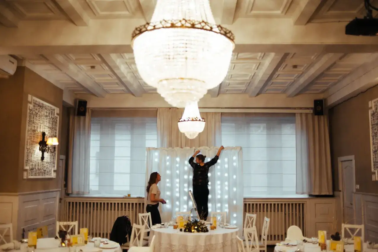 Dwie kobiety przygotowują salę weselną, jedna stoi na drabinie, dekorując tło światełkami. To początek drogi, jak zostać wedding plannerką.