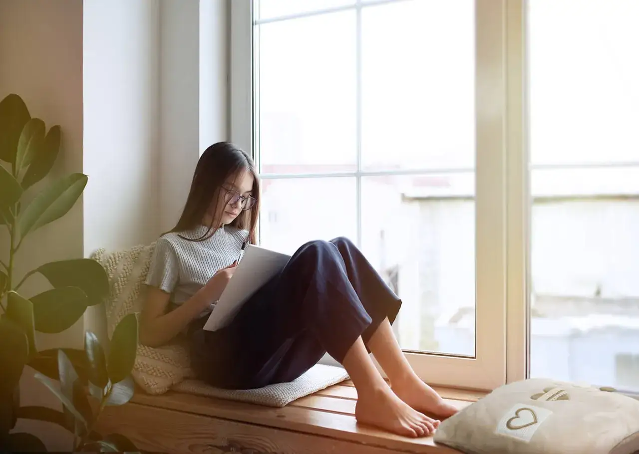 Dziewczynka w okularach siedzi na wygodnym siedzisku pod oknem, rysując coś w szkicowniku.