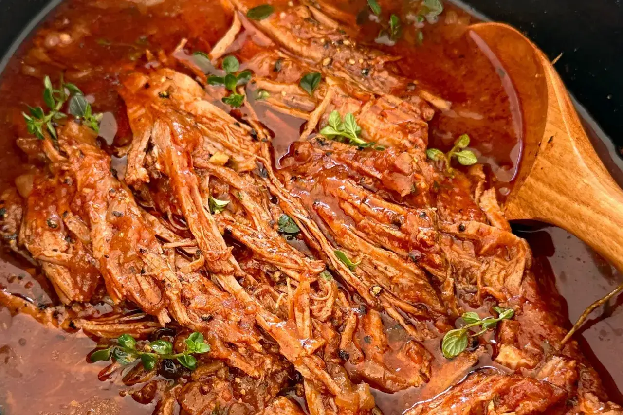 Miękka, szarpana wołowina w sosie pomidorowym, ozdobiona ziołami, obok drewnianej łyżki.