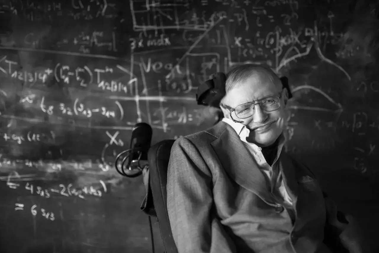 Młody Stephen Hawking w okularach, z uśmiechem, siedzi na wózku inwalidzkim przed tablicą pełną równań.