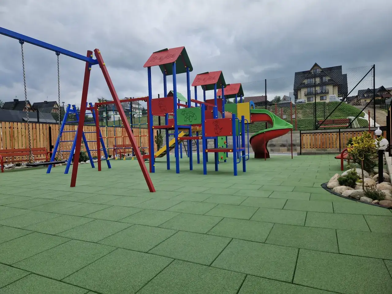 Kolorowy plac zabaw z zieloną, gumową nawierzchnią, huśtawką i zjeżdżalnią.