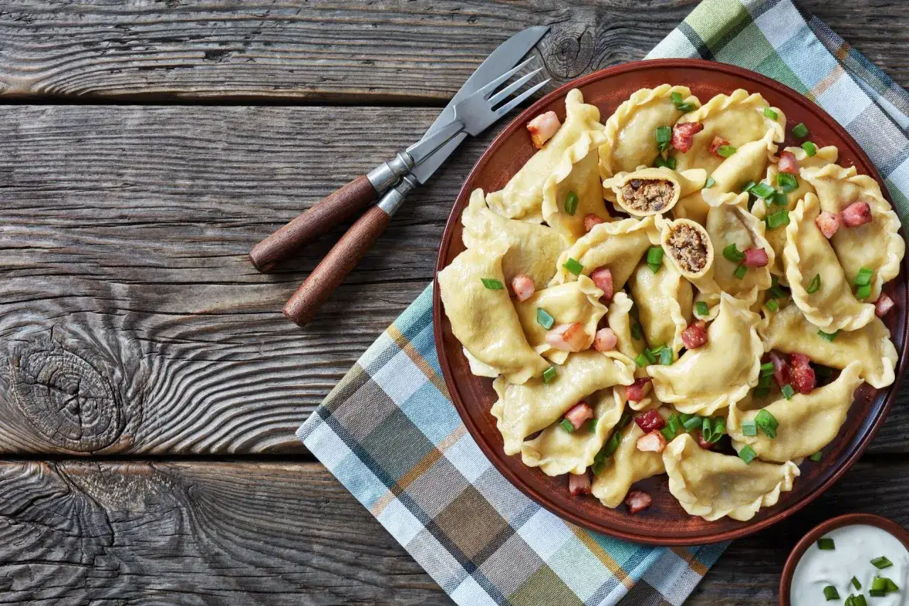 Pyszne pierogi z farszem na pierogi z mięsem mielonym, posypane szczypiorkiem i skwarkami. Obok widelec i nóż.