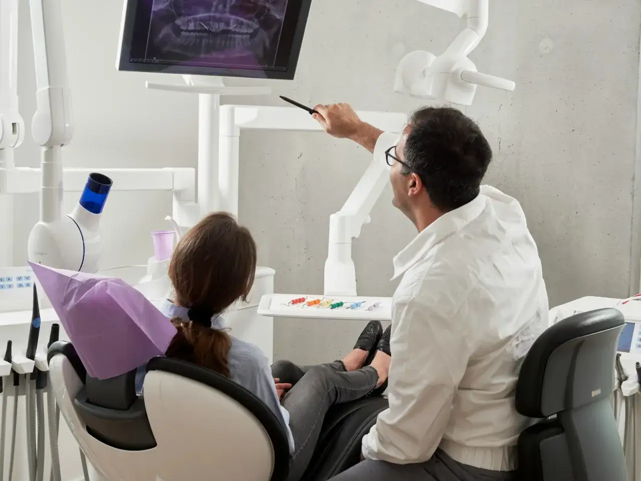 Dentysta w białej koszuli wskazuje na ekran z prześwietleniem zębów pacjentce siedzącej na fotelu.