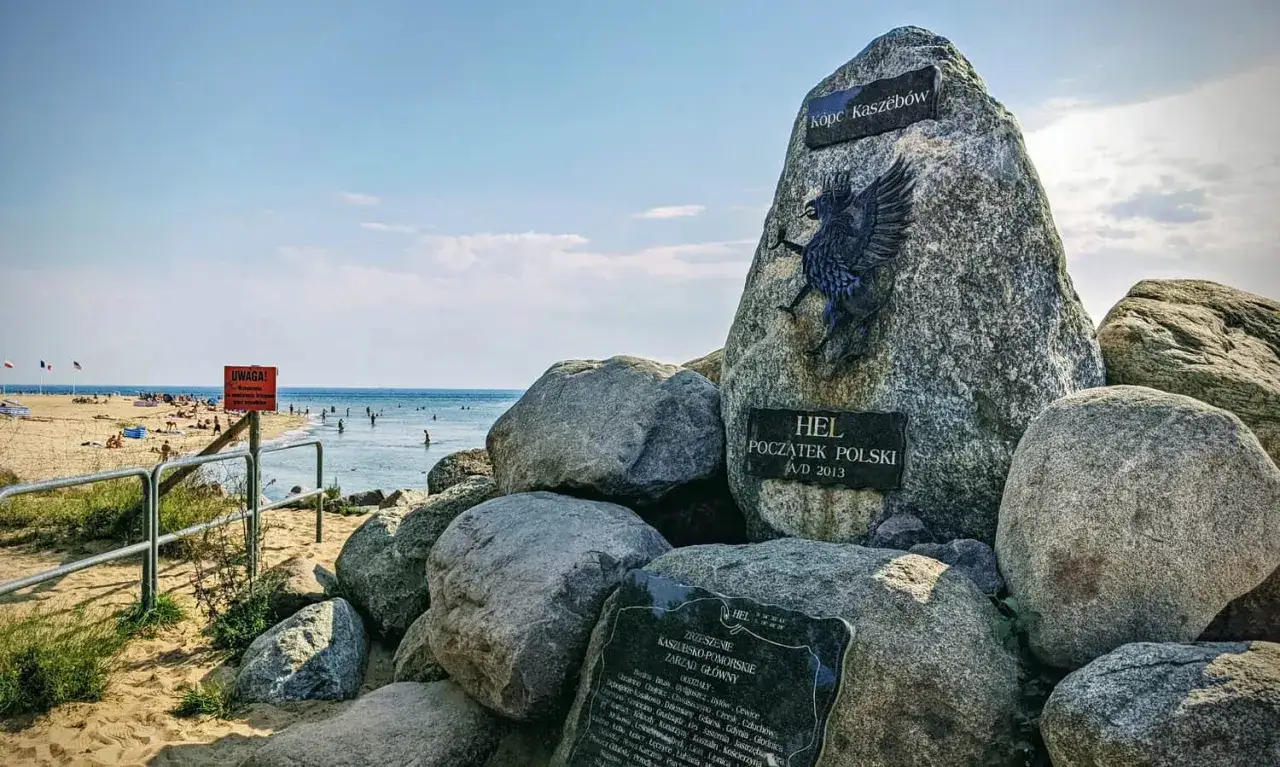 Pomnik "Początek Polski" na plaży na cyplu helskim. W tle widać morze i plażowiczów.