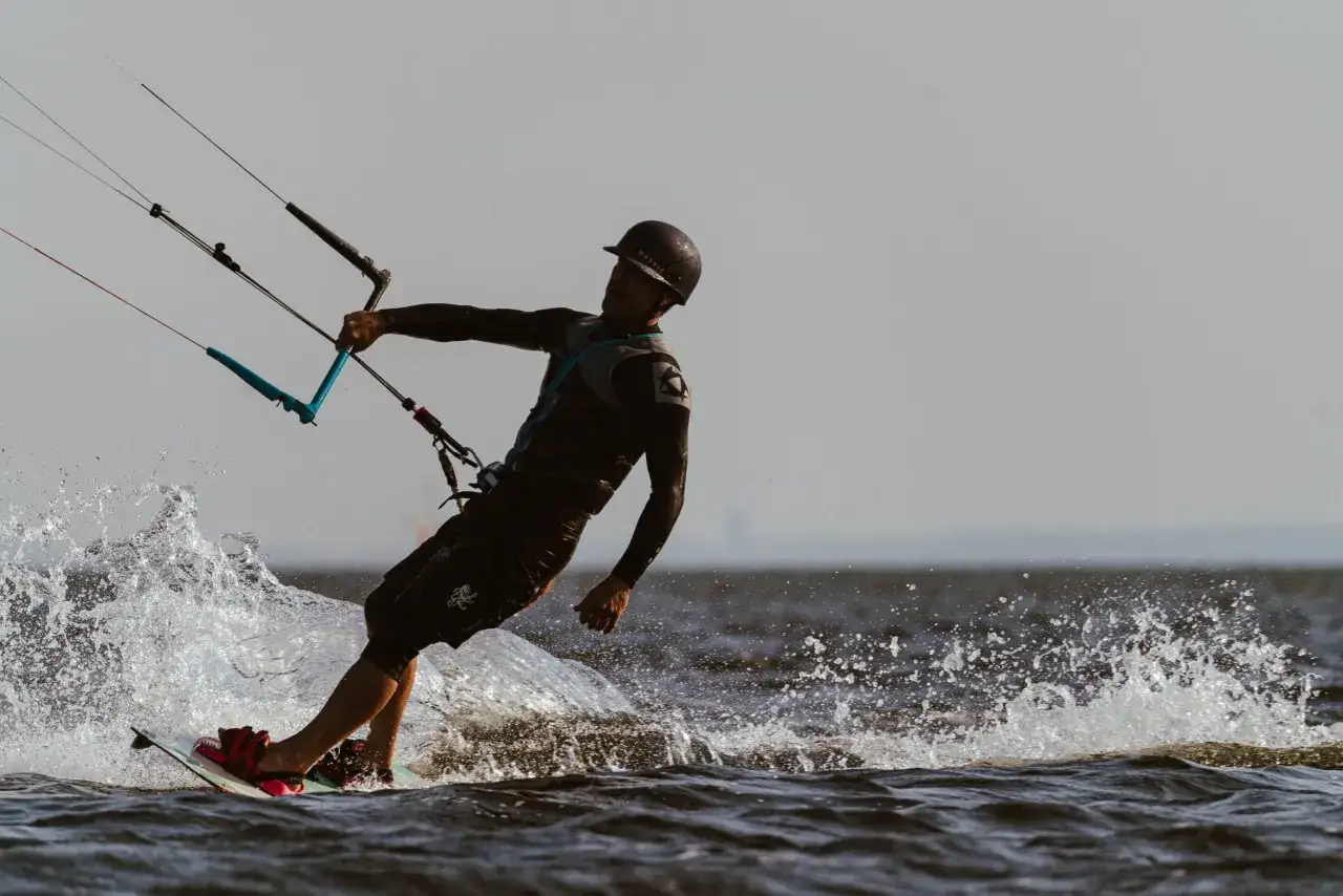 Kitesurfer w kasku i piance, z latawcem i deską, tworzy rozpryski wody na jeziorze.