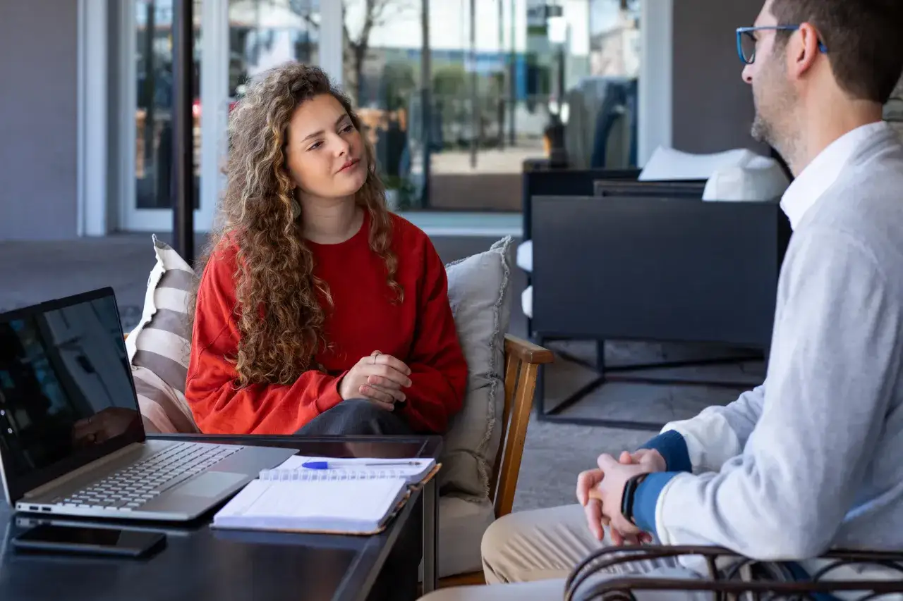Kobieta z kręconymi włosami i mężczyzna rozmawiają przy stoliku z laptopem i notatnikiem.