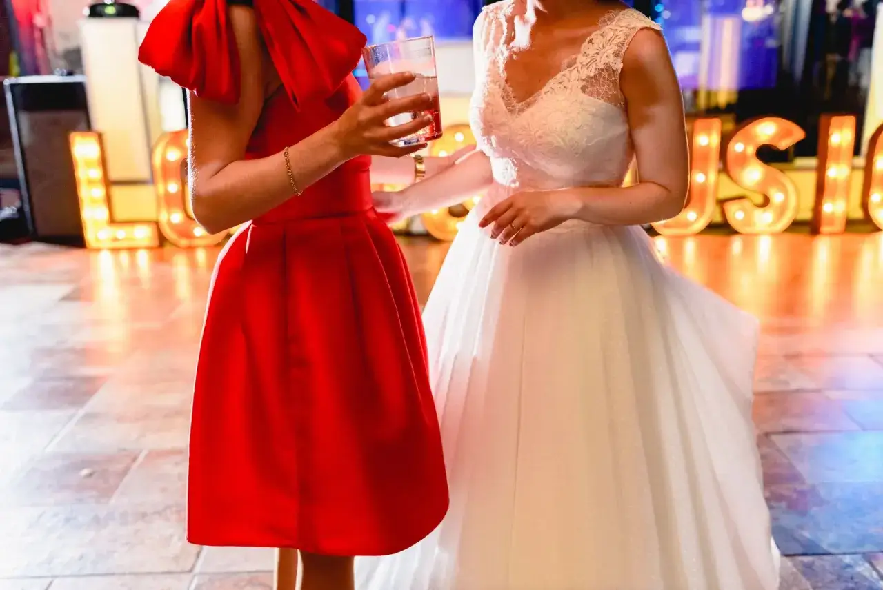 Dwie kobiety w sukniach: jedna czerwona, druga biała, na tle podświetlonych liter.