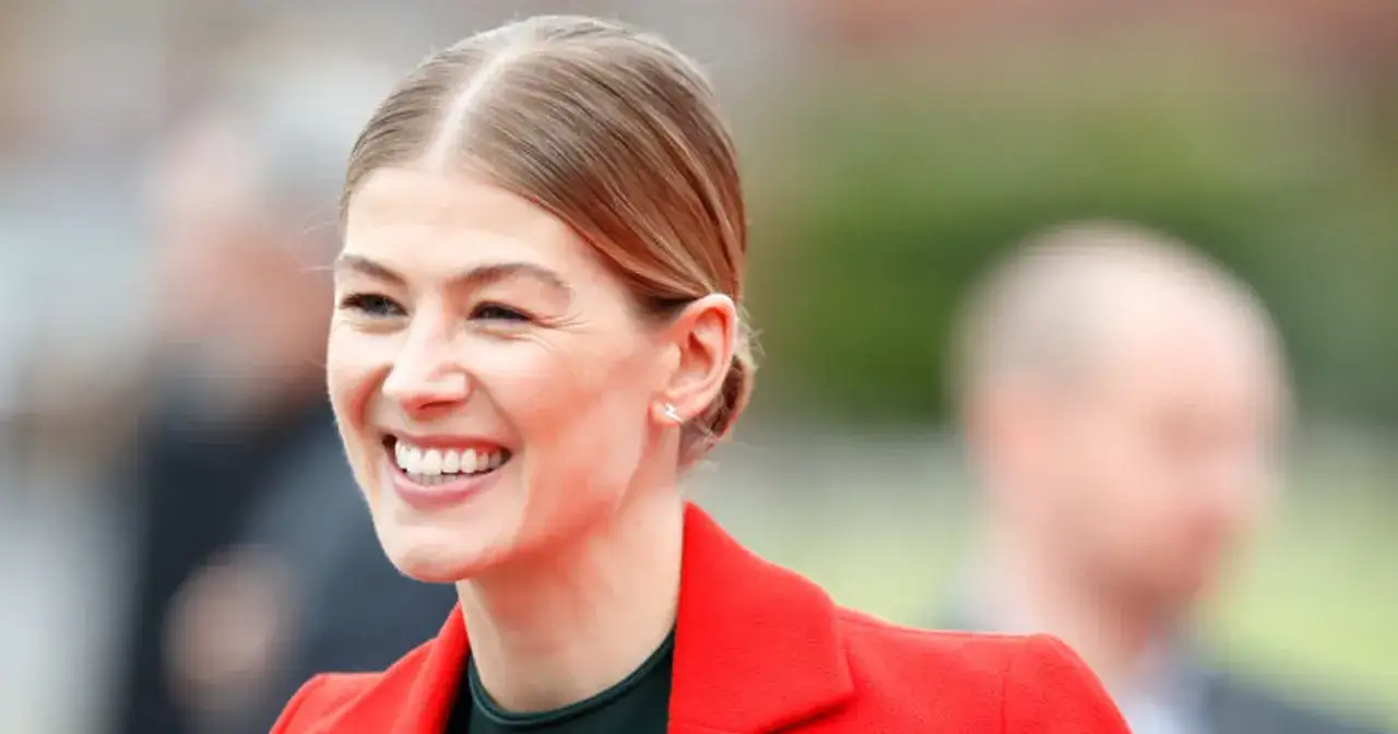 Uśmiechnięta Rosamund Pike, znana z wielu filmów i seriali, w czerwonej marynarce.