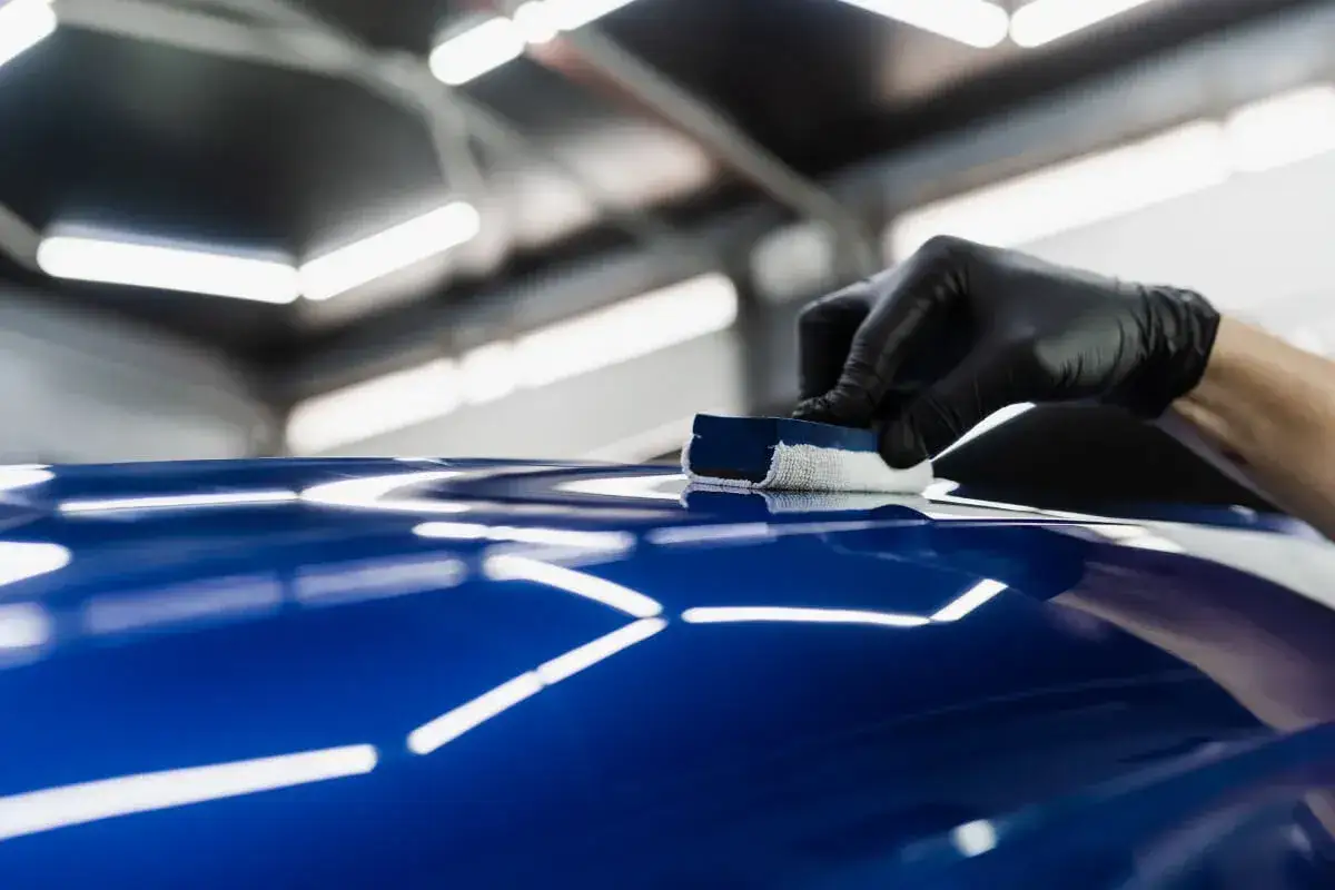 Ręka w czarnej rękawiczce nakłada wosk na błyszczący, granatowy lakier samochodu.