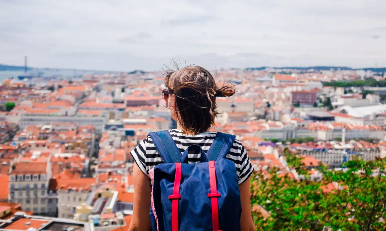 Widok zza pleców osoby z plecakiem na miasto z czerwonymi dachami.