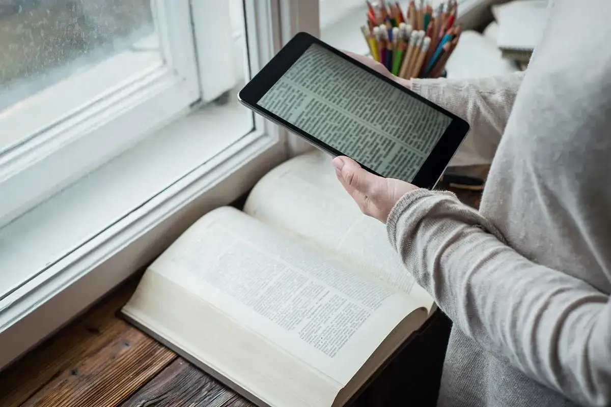 Osoba porównuje tekst na tablecie z otwartą księgą. Czy warto czytać książki? Przykłady z literatury inspirują do poszukiwań.