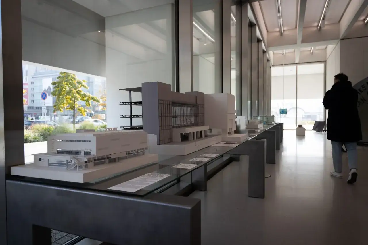 Modele budynków muzealnych na wystawie, z widokiem na miasto przez okno.