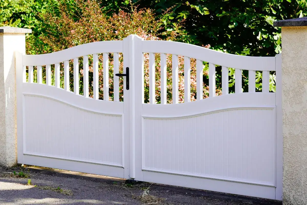 Elegancka, biała brama wjazdowa dwuskrzydłowa z furtką, ozdobiona pionowymi sztachetami i łukowatym wykończeniem.