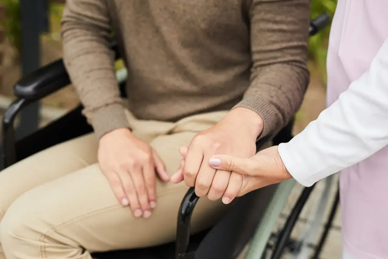 Dłoń opiekuna trzyma dłoń osoby na wózku inwalidzkim.