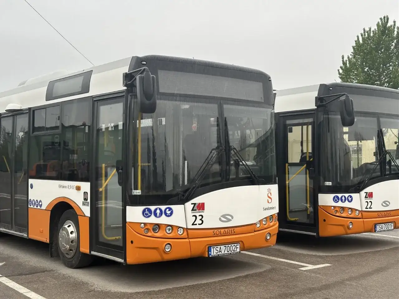 Dwa autobusy miejskie marki Solaris w kolorach białym i pomarańczowym, zaparkowane na placu.