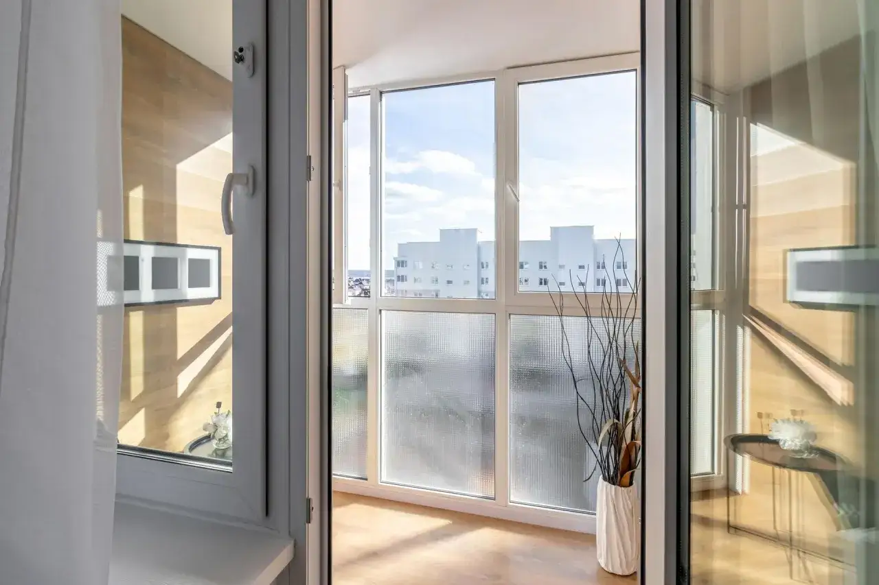 Zabudowany balkon z widokiem na budynki, ozdobiony wazonem z gałązkami.