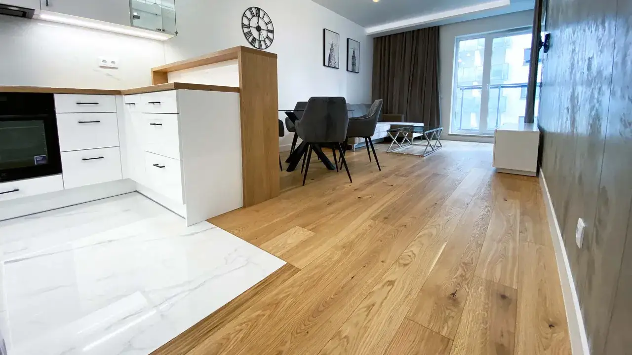 Eleganckie łączenie podłogi drewnianej z płytkami w nowoczesnym wnętrzu. Drewniana podłoga płynnie przechodzi w białe płytki z marmurowym wzorem.