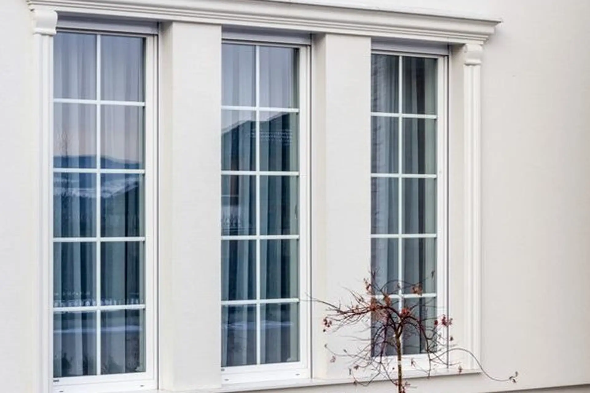 Eleganckie okna z białymi szprosami, tworzące klasyczny podział szyb. Widoczne zasłony i ozdobne obramowanie.