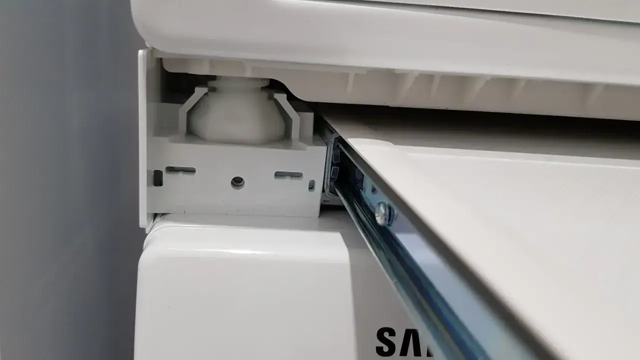 Zbliżenie na mechanizm łącznika suszarki na pralce Samsung, widoczny metalowy wspornik i biała podstawa.