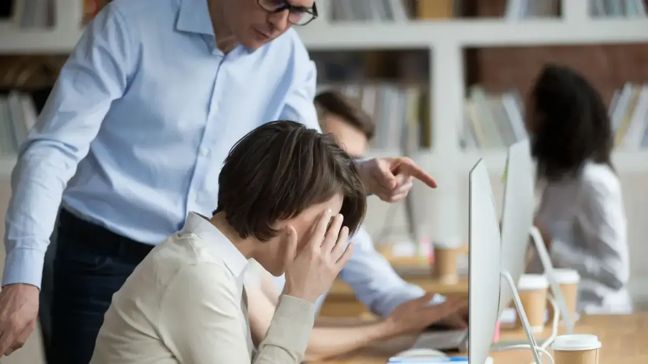 Przełożony wskazuje palcem na pracownicę, która zakrywa twarz dłońmi. Widać niewłaściwe zachowanie przełożonego wobec pracownika.