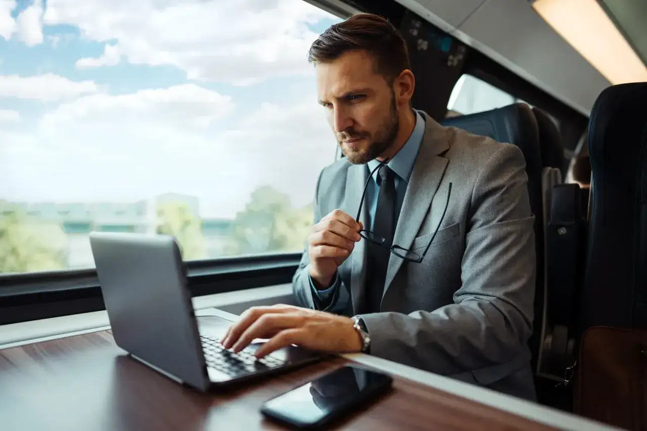 Biznesmen w garniturze pracuje na laptopie w pociągu, trzymając okulary.