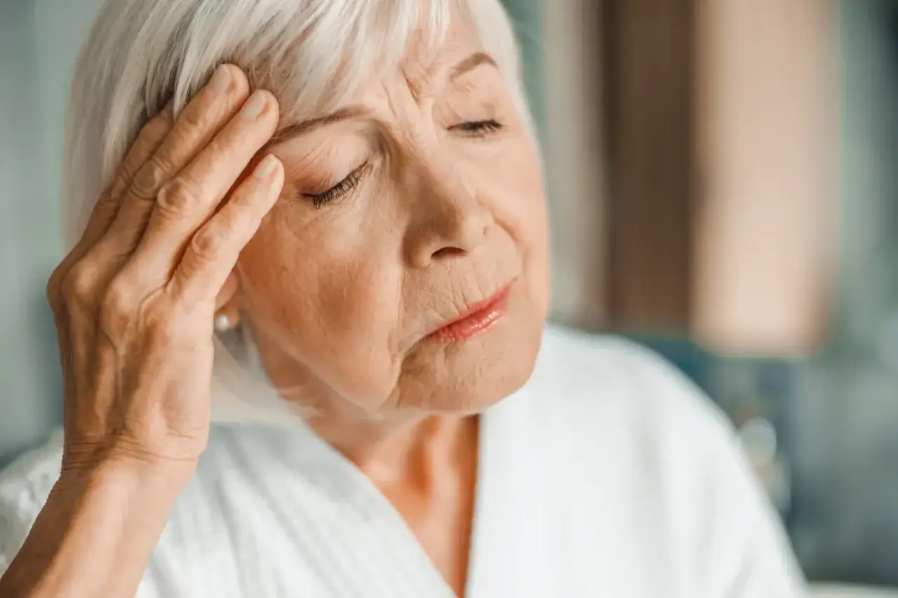 Starsza kobieta z ręką na skroni, zamyślona. Może to być jeden z objawów choroby Alzheimera.