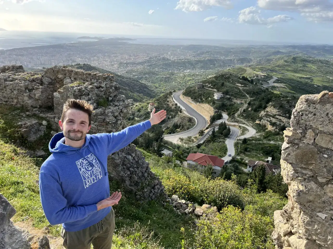 Mężczyzna na tle malowniczego krajobrazu z krętą drogą i miastem w oddali. To piękna Albania zimą, choć zieleń sugeruje wczesną wiosnę.