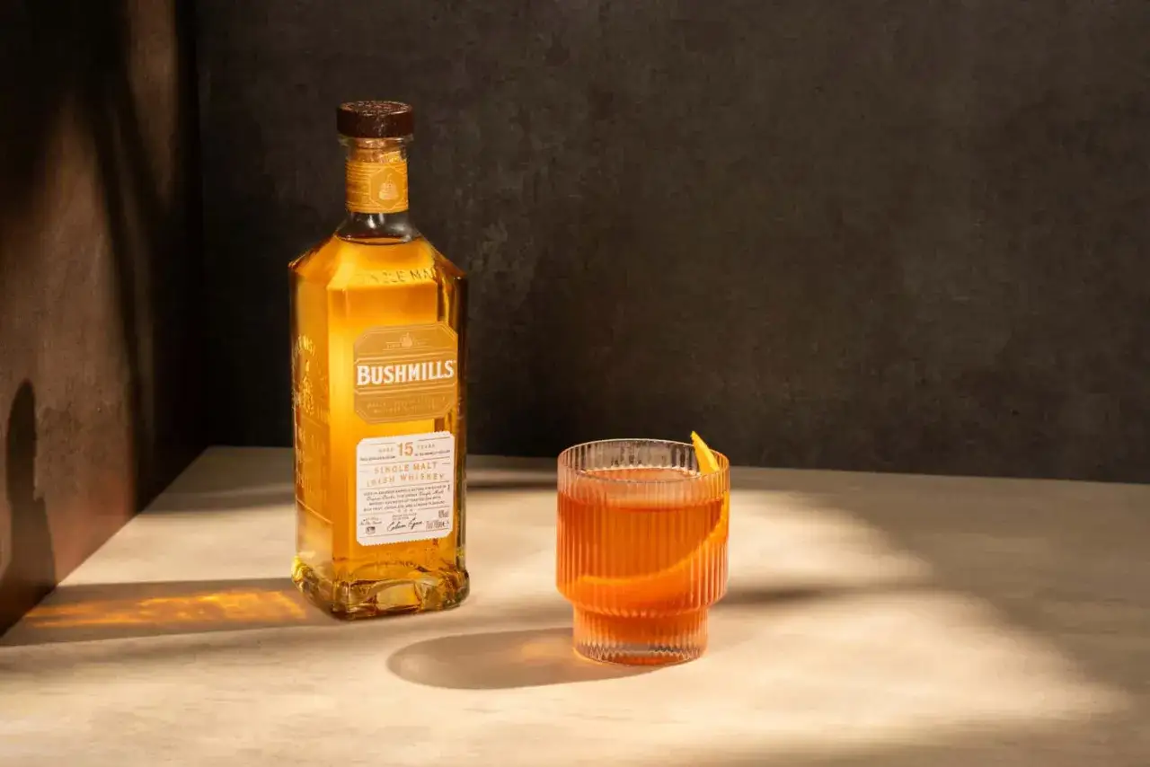 Butelka Bushmills 15 Year Old Single Malt Irish Whiskey obok szklanki z drinkiem. Wiele pozytywnych bushmills whisky opinie potwierdza jego wyjątkowy smak.