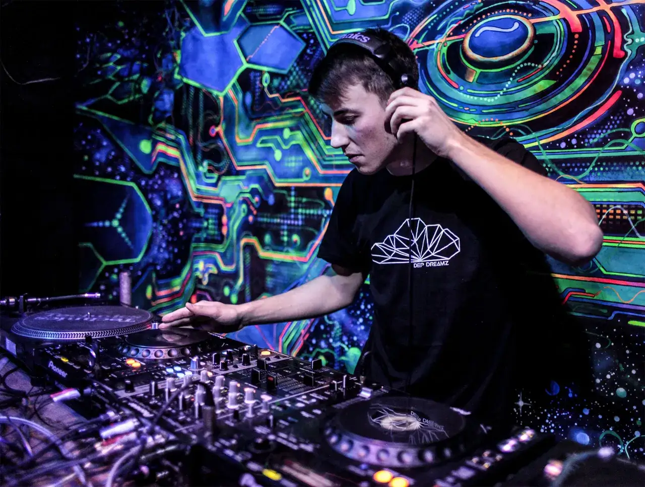Młody DJ w słuchawkach miksuje muzykę na tle psychodelicznej, neonowej ściany.