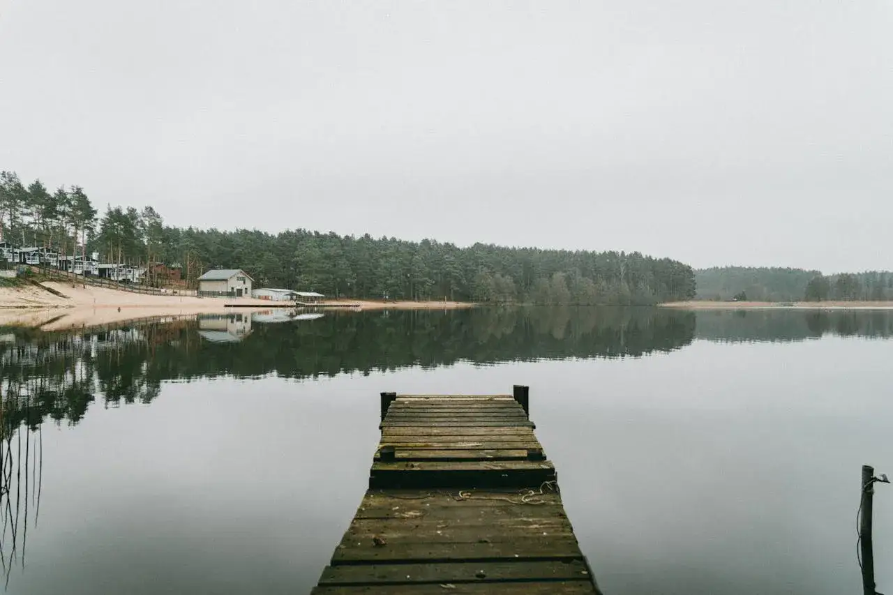 Drewniane molo nad spokojnym jeziorem, z lasem i domkami w tle. To widok, który może skłonić do zastanowienia, ile jest jezior w Olsztynie.