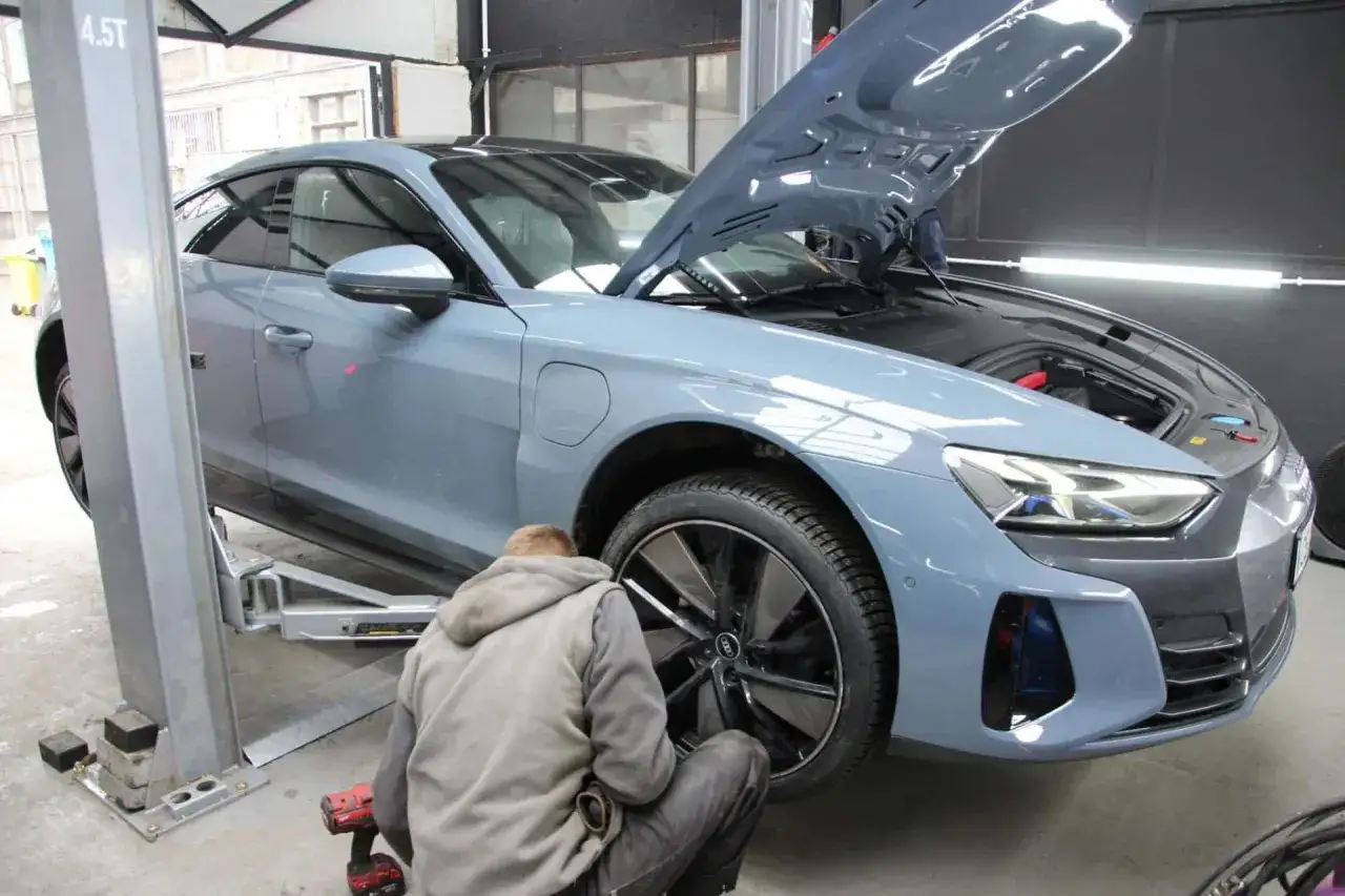 Mechanik przykręca koło niebieskiego samochodu na podnośniku, maska jest otwarta.