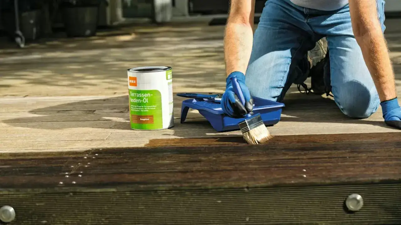 Osoba w niebieskich rękawiczkach nakłada olej na drewniany taras, dbając o jego konserwację.